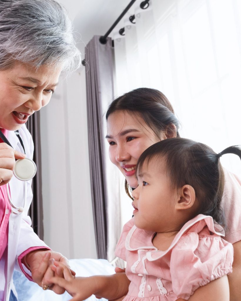 Happy senior female pediatrician professional doctor talk and play to asian girl sitting on mother before exam treatment at hospital or clinic. asia medical child health care.