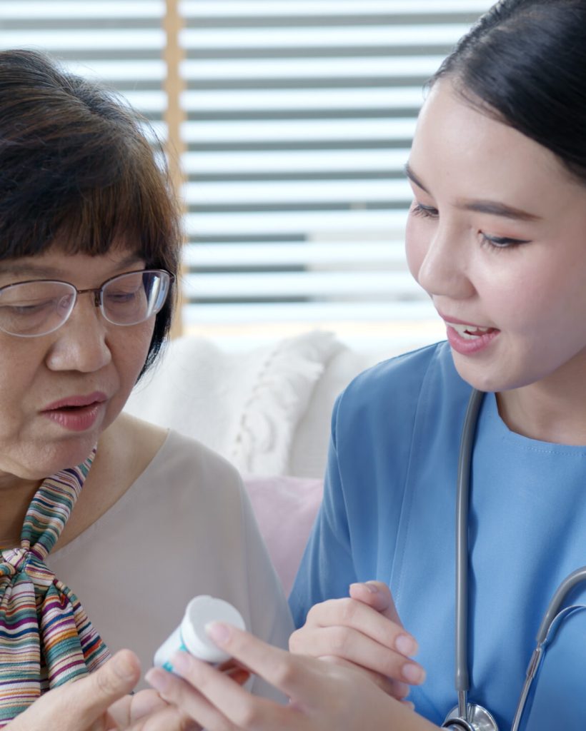 Young caregiver in scrubs uniform showing medicine bottle to old elderly asian woman in home visit care nursing service.  Asian senior with assisted living medication monitoring sit and talk at home.