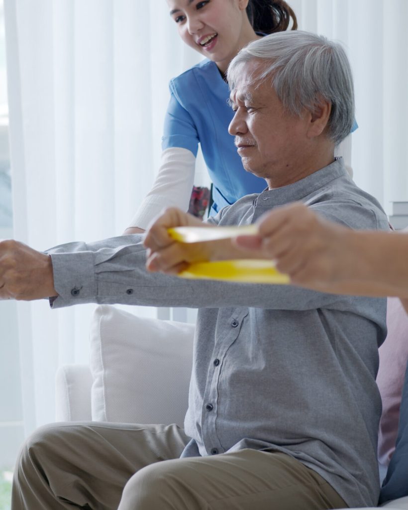 Young older senior asia citizen couple sit on sofa couch with in-home nursing care, assisted living, scrubs nurse use resistance band exercise for senior patient in physiotherapy treatment at home.